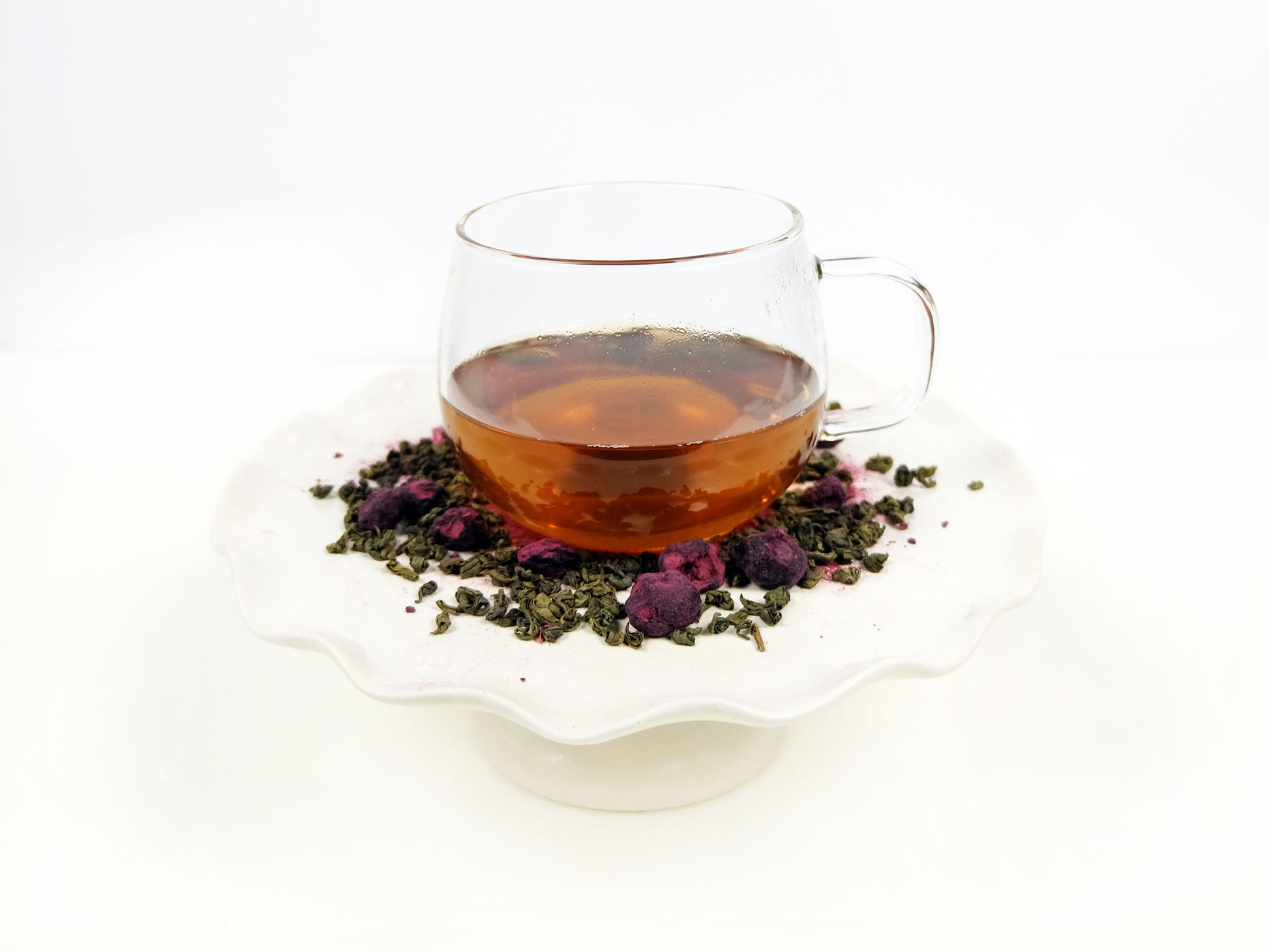 Blueberry Green Tea brewed in a glass mug on a pedestal with loose leaf tea around it
