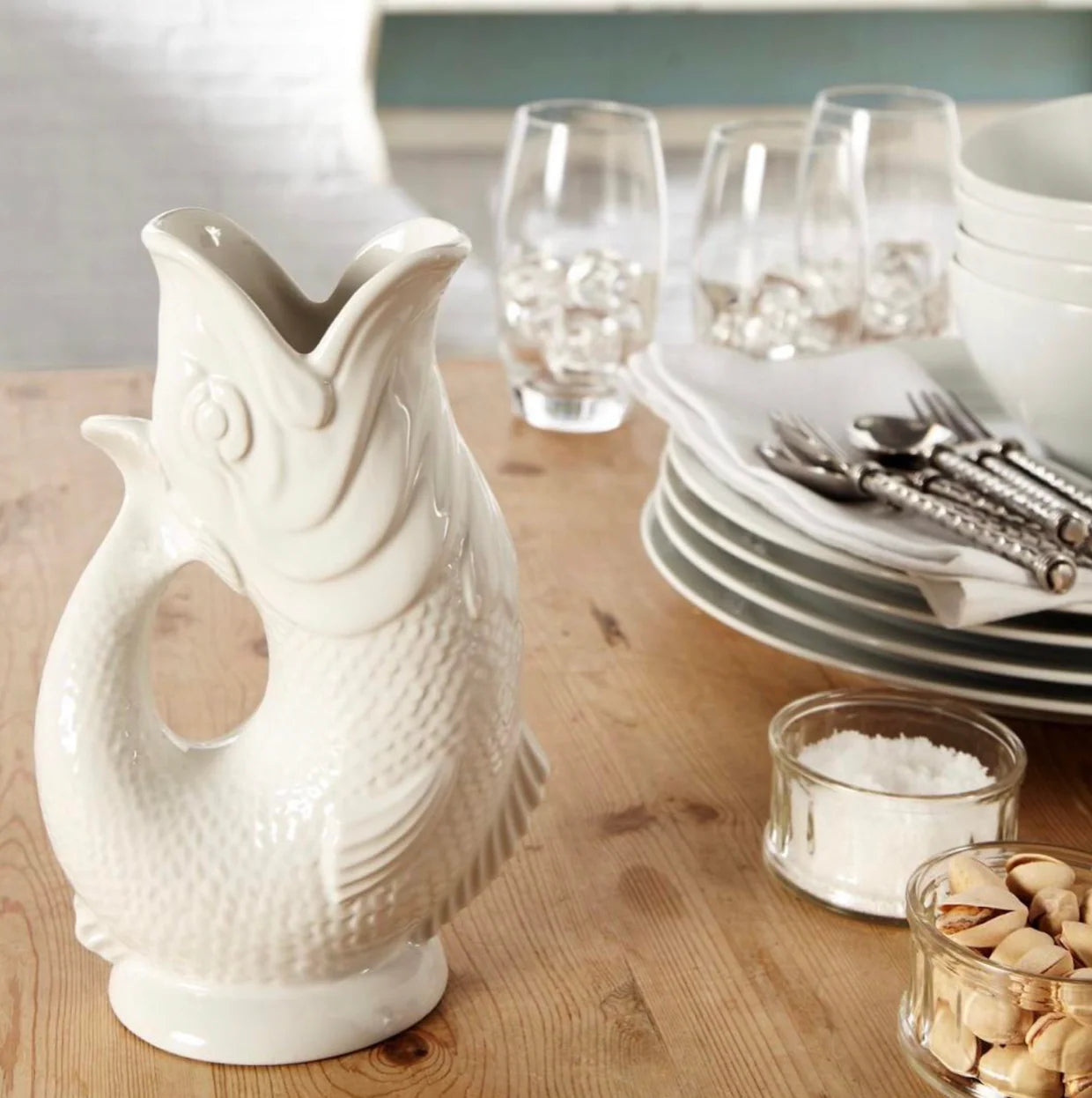 White ceramic fish-shaped gluggle jug on a wooden table with glasses and plates in the background.