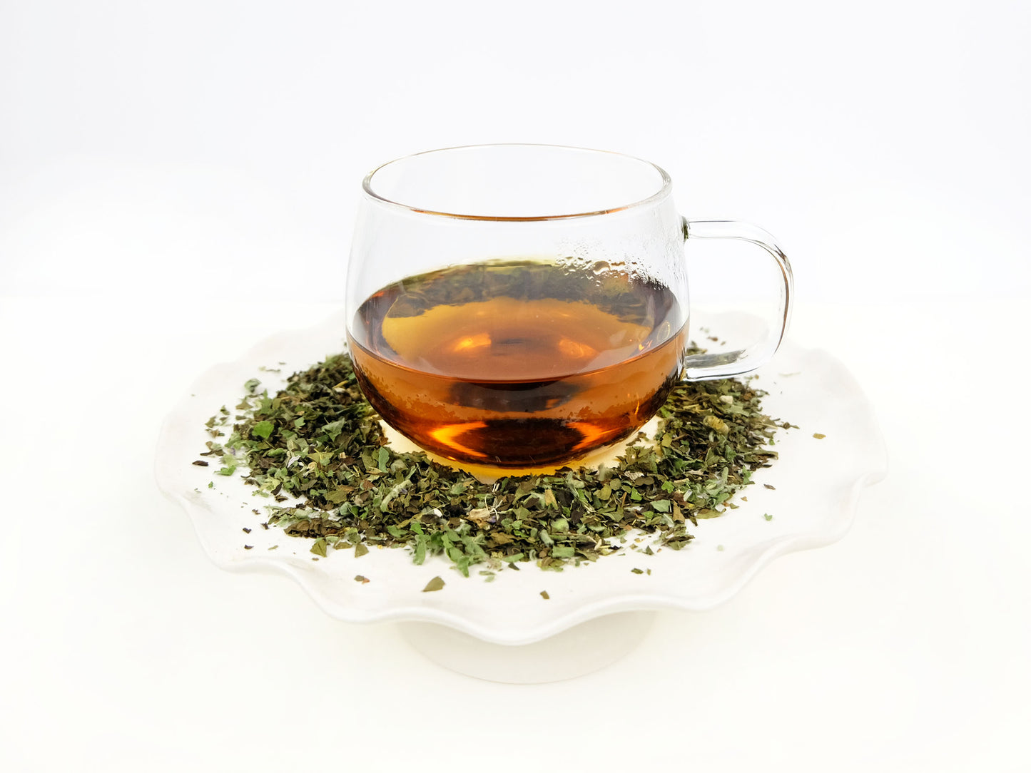 Marshmallow Mint Tea brewed in a glass mug on a pedestal with loose leaf tea around it
