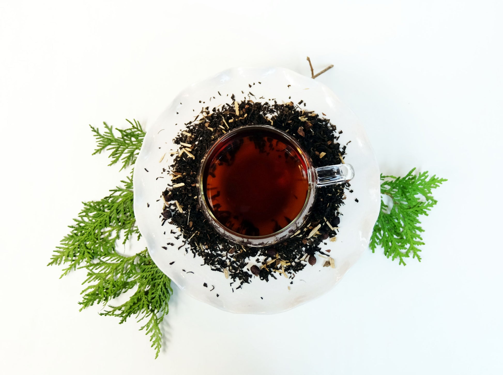 Winter Pine Hawthorn Berry Tea brewed in a glass mug with loose leaf tea scattered around and evergreen branch, top view